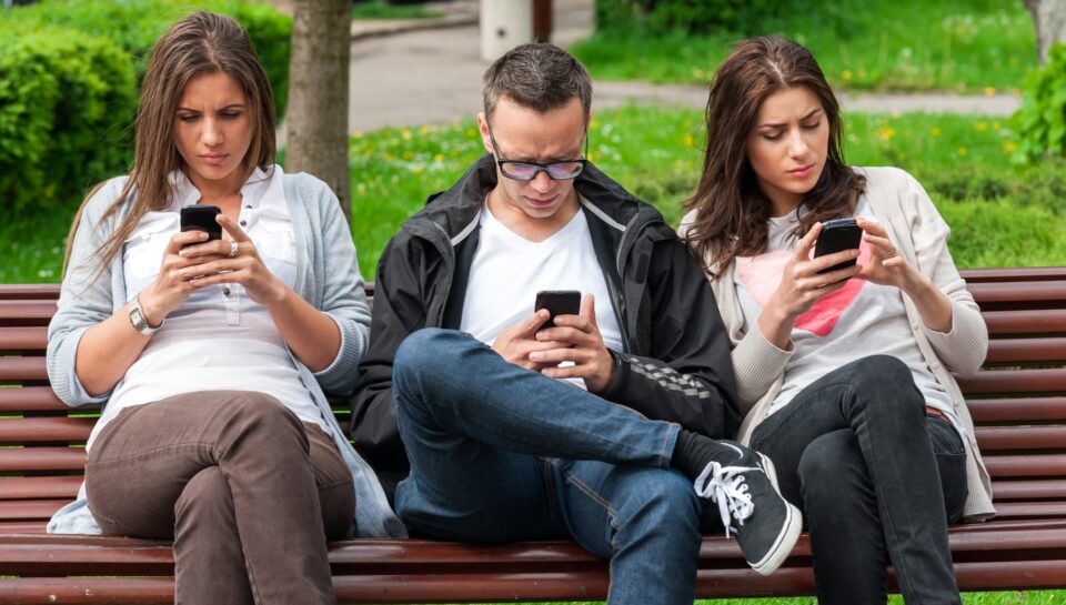 three people on bench looking down at cell phone with tech neck